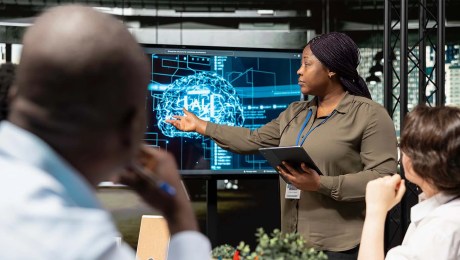 A person presenting to two seated individuals. The presenter is pointing to a digital display with a graphic image of a brain with 'AI' written in the centre.