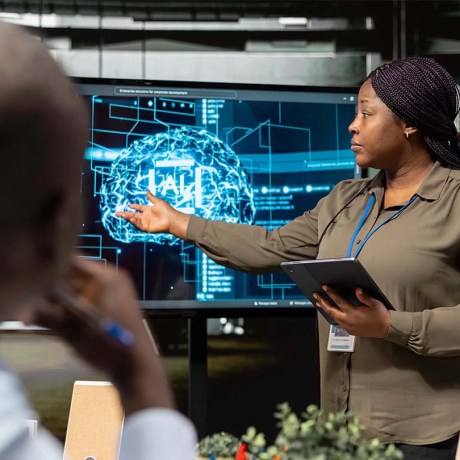 A person presenting to two seated individuals. The presenter is pointing to a digital display with a graphic image of a brain with 'AI' written in the centre.