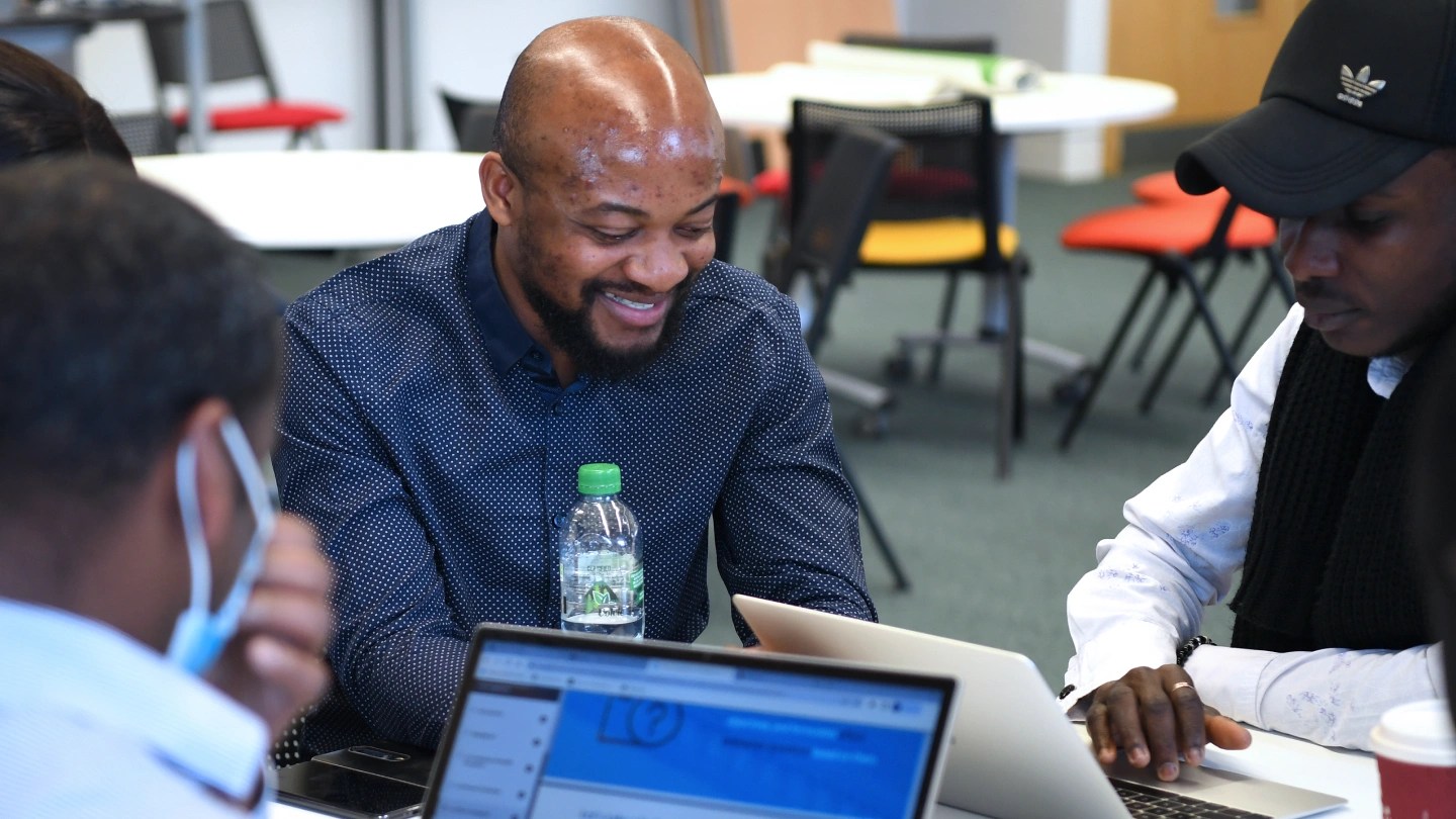 Three people sitting at a table and working on laptops in a classroom.