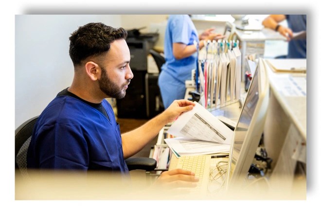 A nurse flipping through papers whilst looking at a computer screen at a hospital reception desk.